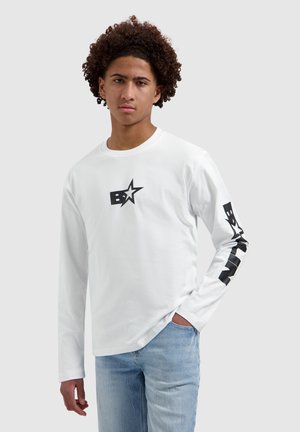 Jonge man met krullend haar die een wit lang mouwen shirt draagt met een sterrenlogo op de borst en zwarte tekst op de mouw, gecombineerd met lichtblauwe spijkerbroek.