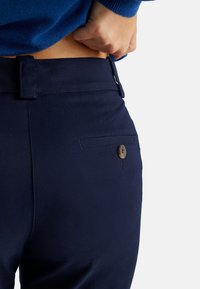 Granatowe spodnie mają gładką teksturę, dopasowany krój oraz tylną kieszeń z okrągłym brązowym guzikiem. Pas znajduje się tuż poniżej klatki piersiowej.