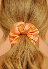 Ein orangefarbener Satin-Haargummi hält das Haar zu einem Pferdeschwanz zusammen; er hat eine leicht glänzende Textur und ein Markenetikett mit einem Logo.