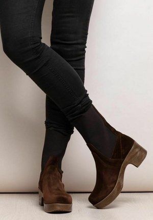 Persoon die zwarte broek en donkerbruine suède plateau enkellaarsjes met houten hakken draagt, staat met één been over het andere gekruist.