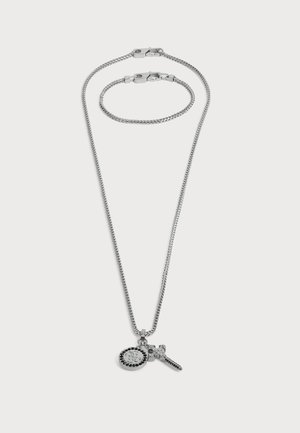 Collar de cadena de plata con colgantes circulares y en forma de cruz, combinado con una pulsera de cadena de plata a juego sobre un fondo blanco.