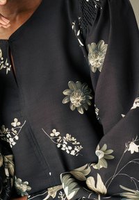 Schwarze Bluse mit floralem Muster, die große graue Blumen und zarte weiße Akzente zeigt, mit einem V-Ausschnitt und strukturierten Ärmel-Details.