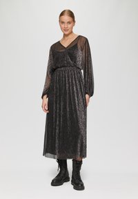 Schwarzes, plissiertes Maxikleid mit transparenten langen Ärmeln und V-Ausschnitt. Aus strukturiertem Stoff mit Schimmer gefertigt. Kombiniert mit schwarzen Kampfstiefeln.