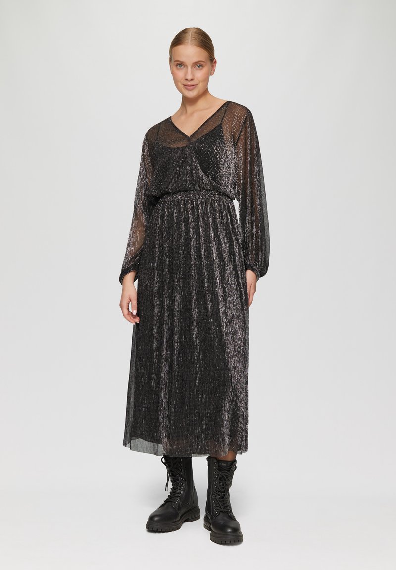 Schwarzes, plissiertes Maxikleid mit transparenten langen Ärmeln und V-Ausschnitt. Aus strukturiertem Stoff mit Schimmer gefertigt. Kombiniert mit schwarzen Kampfstiefeln.