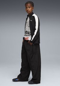 Chaqueta deportiva negra con rayas blancas, camiseta gráfica blanca, pantalones negros holgados y sandalias negras con acentos azules.