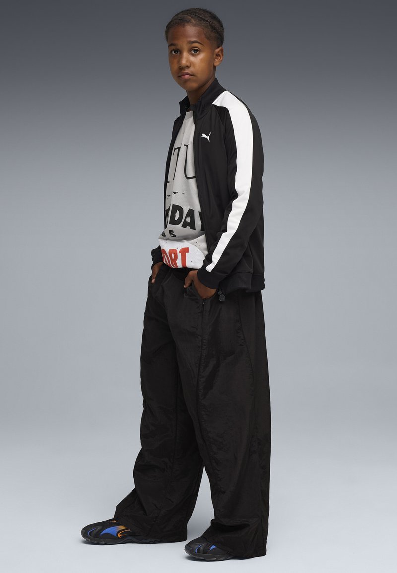 Chaqueta deportiva negra con rayas blancas, camiseta gráfica blanca, pantalones negros holgados y sandalias negras con acentos azules.