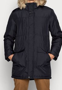 Parka invernale nera con cappuccio foderato in pelliccia, tasche anteriori a patta e bottoni a pressione, indossata sopra pantaloni marroni, mani nelle tasche.