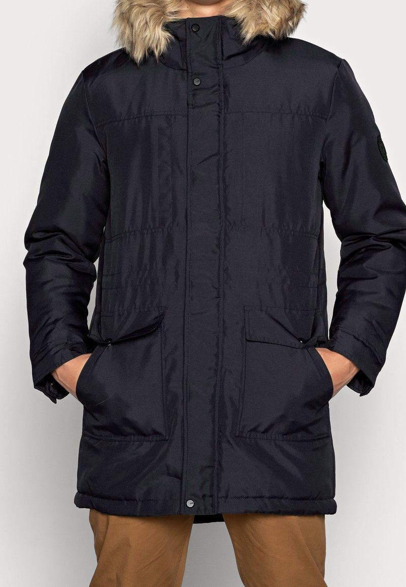 Parka invernale nera con cappuccio foderato in pelliccia, tasche anteriori a patta e bottoni a pressione, indossata sopra pantaloni marroni, mani nelle tasche.