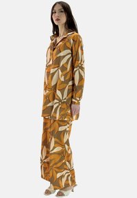 Completo floreale marrone composto da una camicia lunga con bottoni e pantaloni larghi. Il tessuto ha una texture liscia con motivi di foglie beige e arancioni.