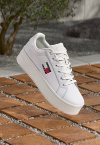Biele platformové tenisky s logom Tommy Jeans, levitujúce nad vonkajším povrchom z dreva a štrku, s zeleňou v pozadí.