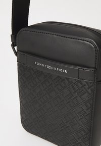 Svart crossbody-veske laget av syntetisk skinn, med et strukturert mønster og et logo-plakett. Inkluderer en justerbar stropp for bæring.