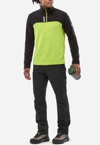 Black and green fleece pullover with a high collar and quarter zipper, paired with black pants and grey hiking boots, holding a green water bottle.