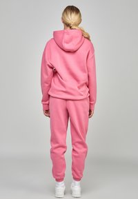 Felpa rosa con pantaloni coordinati in tessuto morbido. La felpa ha un cordoncino e una tasca a marsupio, mentre i pantaloni presentano polsini elasticizzati.