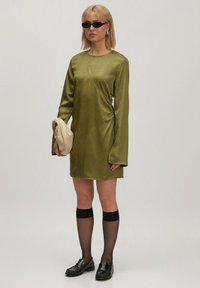 Olijfgroen satijnen jurk met lange mouwen en gerimpelde zijdetails. Zwarte kniehoge sokken en zwarte loafers, terwijl ze een beige clutch tas vasthoudt.