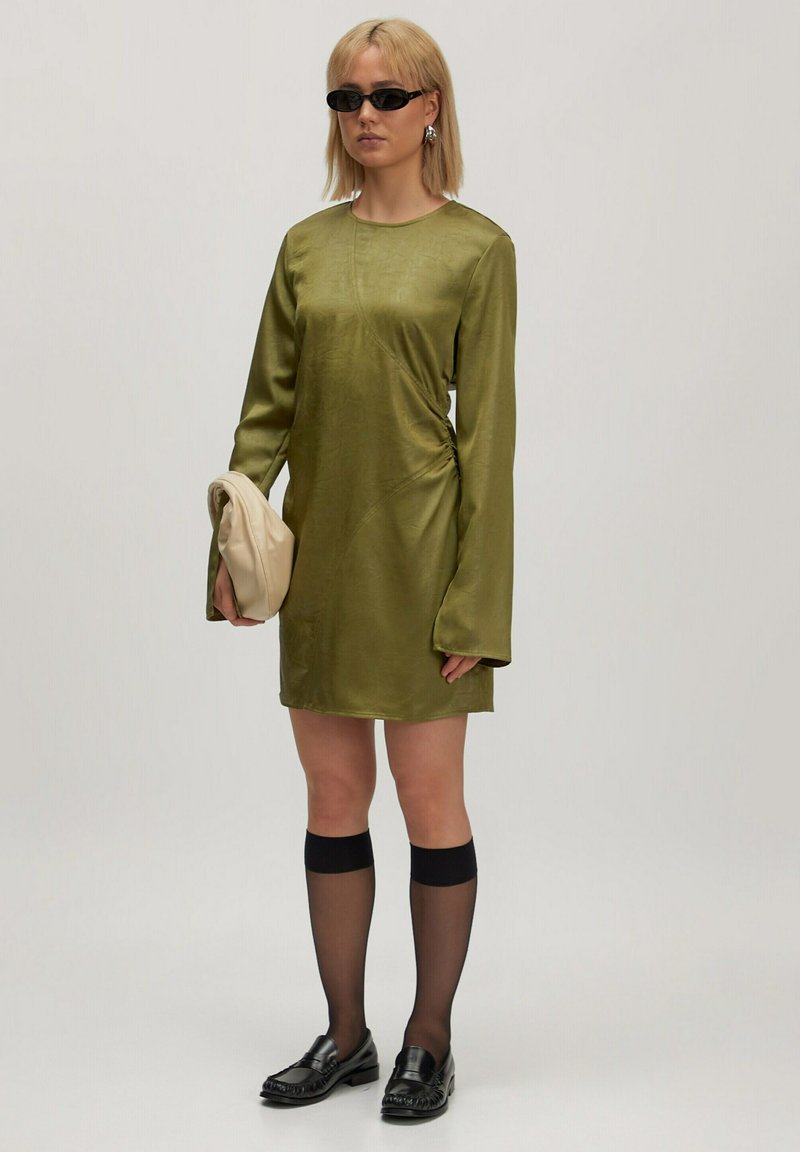 Olijfgroen satijnen jurk met lange mouwen en gerimpelde zijdetails. Zwarte kniehoge sokken en zwarte loafers, terwijl ze een beige clutch tas vasthoudt.