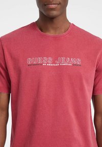 Vyras vilkintis nudėvėtą raudoną apvalios kaklo marškinėlį su užrašu "GUESS JEANS autentiška amerikietiška tradicija džinsai" atspausdintu balta ir juoda spalvomis.