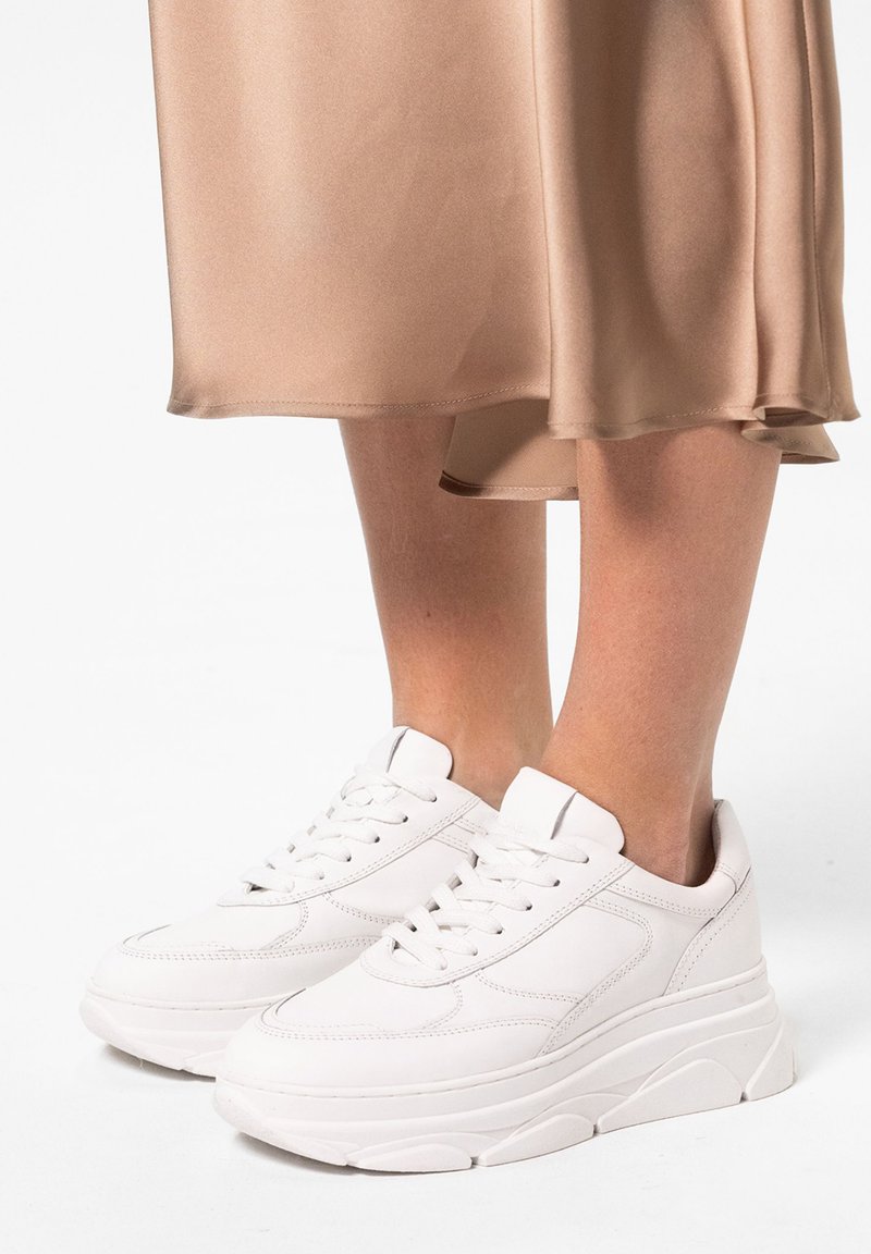 Weißer Sneaker aus glattem Leder, ausgestattet mit einer dicken Sohle, runder Zehenpartie und klassischem Schnürdesign, kombiniert mit einem satinbeigen Rock.