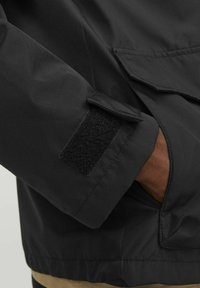Black jacket featuring a Velcro closure on the cuff, with smooth texture and a boxy fit. Includes a side pocket with flap detail.