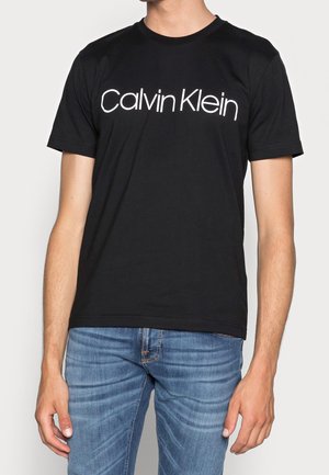 Muž v černém tričku Calvin Klein a modrých džínách stojí s pažemi volně podél těla před jednobarevným pozadím.