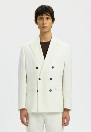 Giovane uomo che indossa una giacca doppiopetto bianca con bottoni neri sopra una camicia nera, in piedi davanti a uno sfondo chiaro semplice.