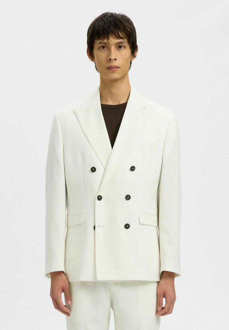 Jeune homme portant une veste de costume croisée blanche à boutons noirs sur une chemise noire, debout devant un fond clair uni.