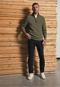 Sudadera verde oliva con cremallera, pantalones negros y zapatillas blancas, con un fondo de madera que presenta listones horizontales.