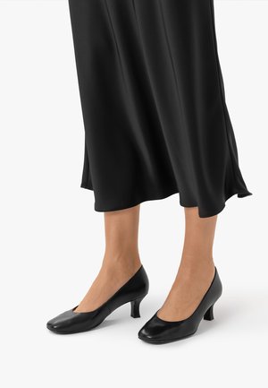Osoba nosi czarne buty na średnim obcasie oraz czarną satynową spódnicę sięgającą poniżej łydek na białym tle.