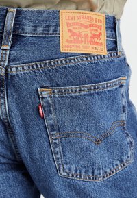 Blå denimjeans med en läderlapp märkt "Levi Strauss & Co." vid midjan, och gula stygn som framhäver fickorna.