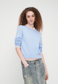 Γυναίκα με σγουρά κόκκινα μαλλιά που φοράει ανοιχτό μπλε πουλόβερ και γκρι τζιν φούστα με σχέδιο από φίδι, με τα χέρια στις τσέπες.