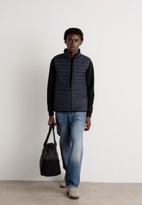 Colete puffer preto com costuras horizontais, usado sobre uma camisola preta de manga longa, combinado com jeans azul claro e um saco preto.