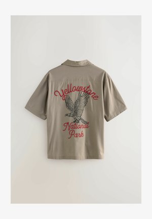 Chemise kaki à manches courtes ornée d'une broderie détaillée d'un oiseau et du texte "Parc national de Yellowstone" en rouge. Texture lisse.