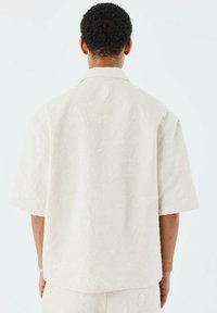 Korte mouw witte blouse met subtiel textuurpatroon, relaxte pasvorm en een puntige kraag. Heeft een rond logo op de bovenrug.