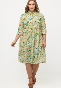 Vestido con cuello floral en verde pastel, que presenta un diseño en capas, mangas de tres cuartos y vibrantes patrones de flores. Sandalias marrones complementan el look.