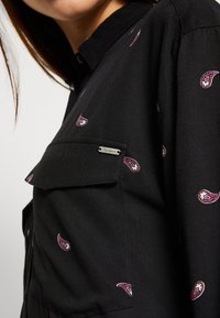 Svart skjorta med kort krage som har små paisleymönster i lila och vitt. Inkluderar en ficka med en silverfärgad märkesskylt.