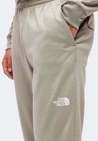 Άτομο που φοράει μπεζ φόρμα The North Face με το ένα χέρι στην τσέπη και με ορατό λογότυπο στο μπούτι.