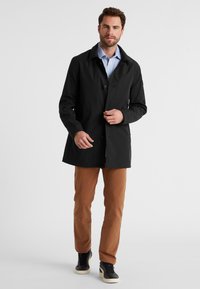 Schwarzer wasserfester Trenchcoat mit geradem Schnitt, Knopfverschluss und Reverskragen; kombiniert mit braunen Hosen und schwarzen Sneakers.