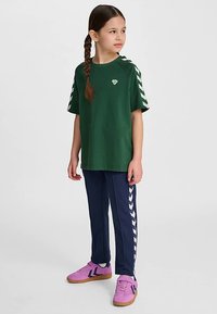 Grön t-shirt med korta ärmar och en liten logga, kombinerad med marinblå byxor med vita chevronränder och rosa sportskor.