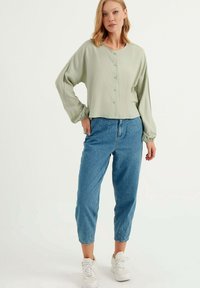 Blusa verde chiaro a maniche lunghe con bottoni e polsini elastici, abbinata a pantaloni cropped in denim blu casual e sneakers bianche.