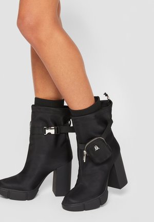Black ankle boots with a glossy finish, featuring a thick block heel, zippered pocket on the side, and a textured knit collar.