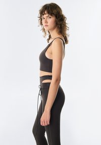 Brązowy stanik sportowy z okrągłym dekoltem i cienkimi ramiączkami, w zestawie z legginsami z wysokim stanem, które mają wycięcia i regulowany sznurek.