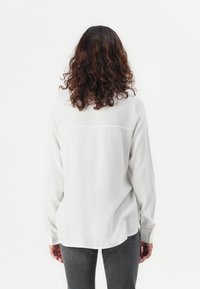 Blusa bianca a maniche lunghe con una texture liscia, cuciture sottili sulla schiena e un orlo curvo, abbinata a pantaloni grigi.