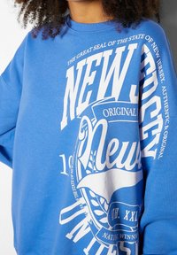 Modrá mikina s velkým bílým textem "New Jersey," "Original" a "Velký státní znak státu New Jersey," kterou nosí osoba.