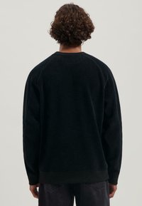 Persona con cabello rizado que lleva un sudadera negra de manga larga lisa y jeans oscuros, mostrada de espaldas contra un fondo neutro.