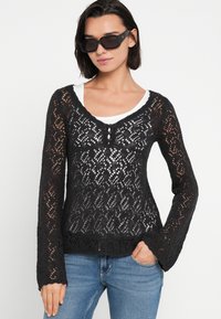 Μαύρο πλεκτό πουλόβερ με μακριά μανίκια και V-λαιμόκοψη, διακοσμημένο με δαντελένια σχέδια και τρία κουμπιά στο λαιμό. Συνδυάζεται με λευκό τοπ.