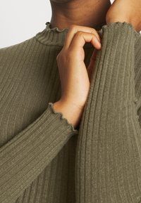 Oliivinvihreä ribbiknitti toppi, jossa on aaltoileva kaulus ja hihansuut, esittelee teksturoitua kangasta ja sukkia muotoa. Käsi pidättelee hihaa.