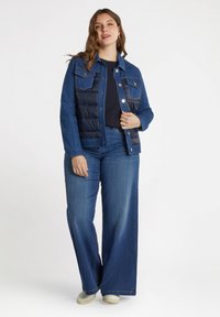 Chaqueta de mezclilla con un panel acolchado azul marino oscuro, con dos bolsillos en el pecho, combinada con jeans de pierna ancha y zapatos de color claro.