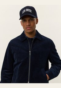 Navy Corduroyjacket mit Reißverschluss und zwei Fronttaschen, über einem gemusterten Hemd getragen. Cap mit "LES DEUX"-Schriftzug in Weiß.