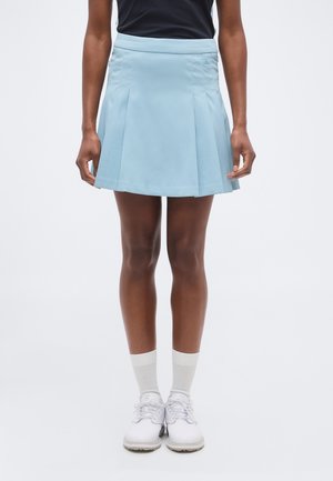 Inimene kannab helehallikat volditud seelikut, musta toppi, valgeid sokke ja valgeid paeltega kingi, seisab ühevärvilise tausta ees.