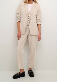 Veston beige ajusté porté sur un gilet et un pantalon assortis, le tout dans un tissu lisse. Mocassins noirs avec des accents de quincaillerie dorée.