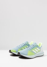 Scarpe da ginnastica azzurro chiaro con accenti verde neon, upper in rete testurizzata, strisce bianche e suola in gomma nera.